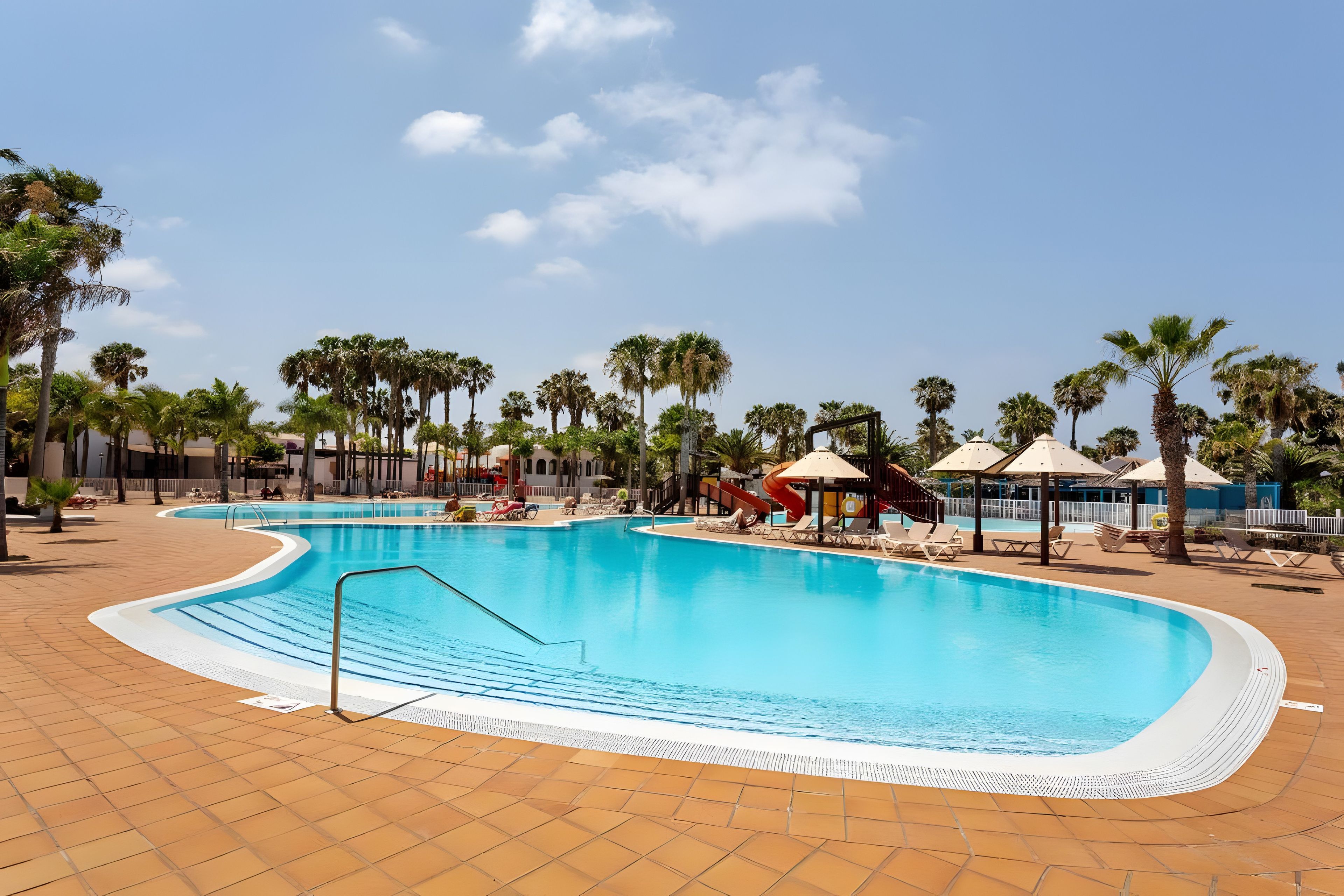 Ein Swimmingpool mit Wasserrutsche, umgeben von Palmen und Liegestühlen.
