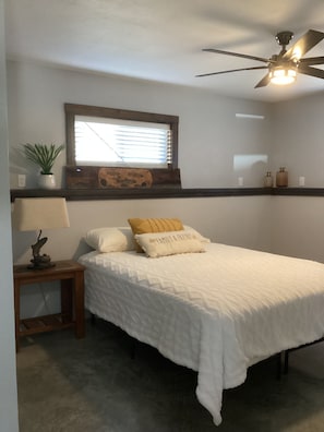 Ein Schlafzimmer mit Bett, Nachttisch, Lampe, Deckenventilator, Fenster mit Jalousien und einem Wandregal mit Dekoration.