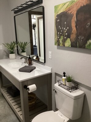 Ein Badezimmer mit einem weißen Waschbecken, Spiegel und Toilette. Auf der Waschtischplatte stehen zwei Zimmerpflanzen und an der Wand hängt ein Bison-Gemälde.