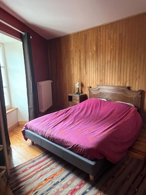 Quarto com cama, mesa de cabeceira, abajur, radiador e janela com cortinas.