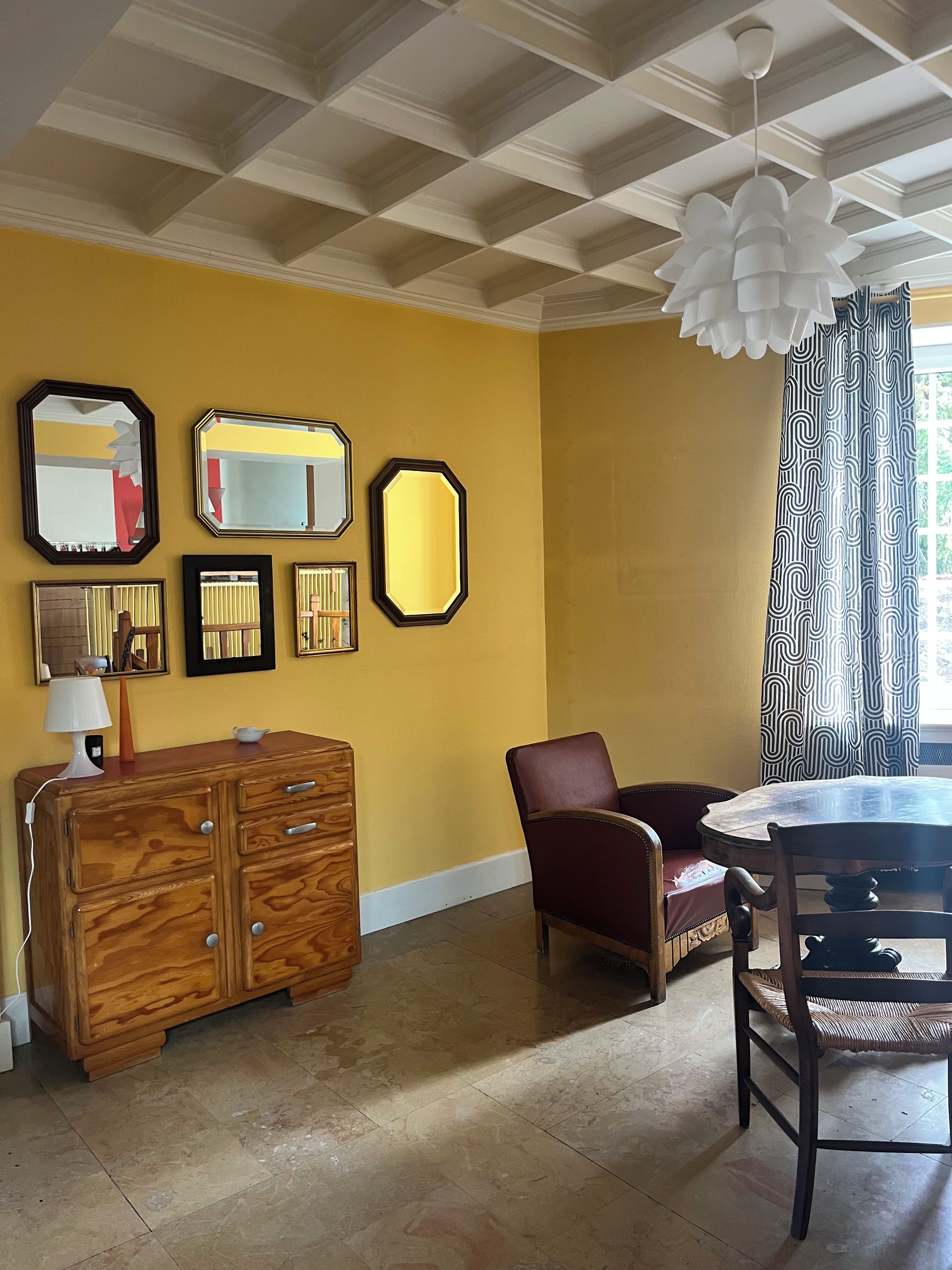 A room with a wooden dresser, a red chair, a dining table with chairs, and a ceiling with a grid pattern.