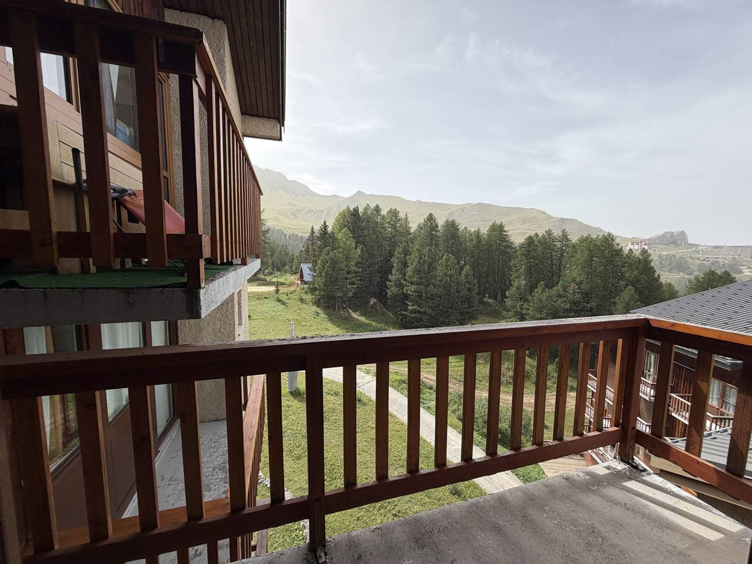 Een balkon met houten hekwerk met uitzicht op een bergachtig landschap met bomen en een gebouw in de verte.