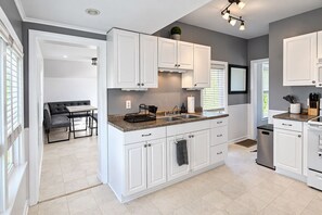Cocina moderna con armarios blancos, encimera de granito y un comedor con sofá gris y mesa.