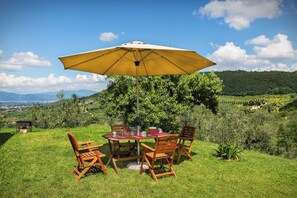 Un área de comedor al aire libre con mesa y sillas bajo un paraguas grande, rodeada de exuberante vegetación y una vista panorámica de un valle y montañas distantes.