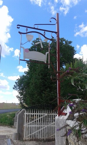 Un letrero de metal con símbolos de cubiertos, una cerca blanca de estacas y un árbol al fondo.