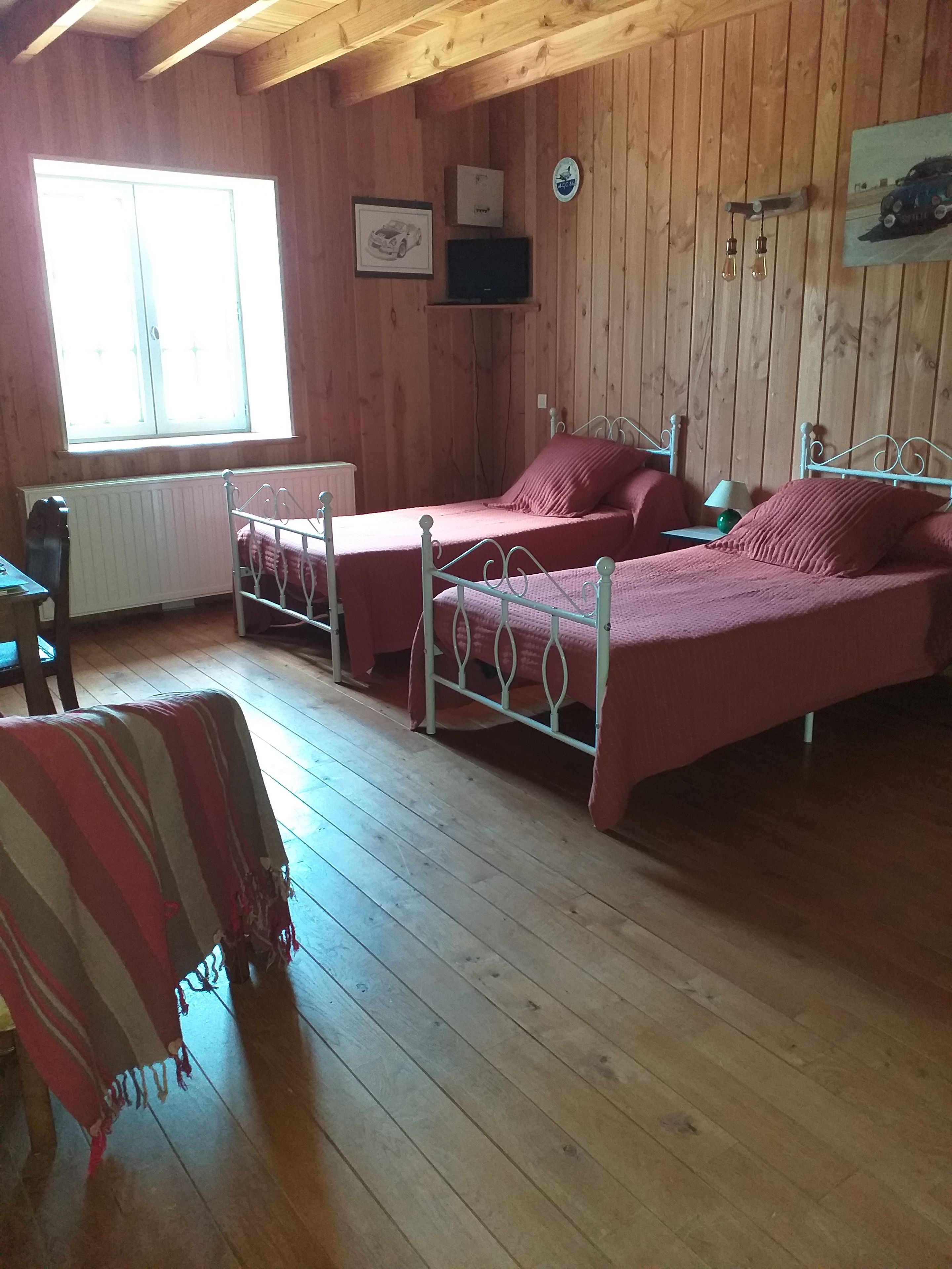 A room with two metal beds, wooden walls, a radiator, a window, and a framed picture on the wall.