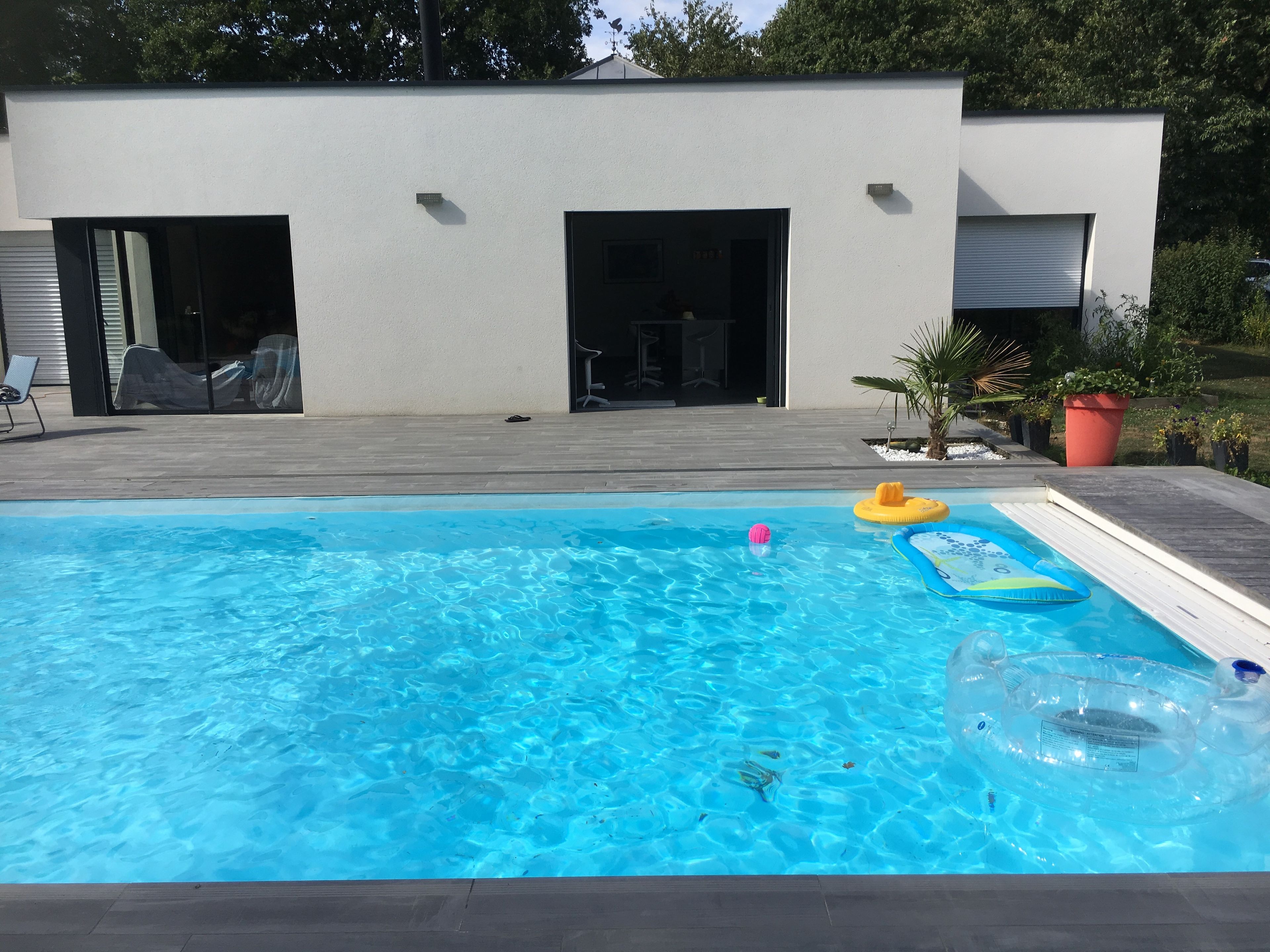 Une piscine avec des jeux gonflables, un patio couvert et un bâtiment moderne aux portes ouvertes.