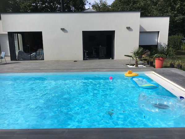 Une piscine avec des jeux gonflables, un patio couvert et un bâtiment moderne aux portes ouvertes.