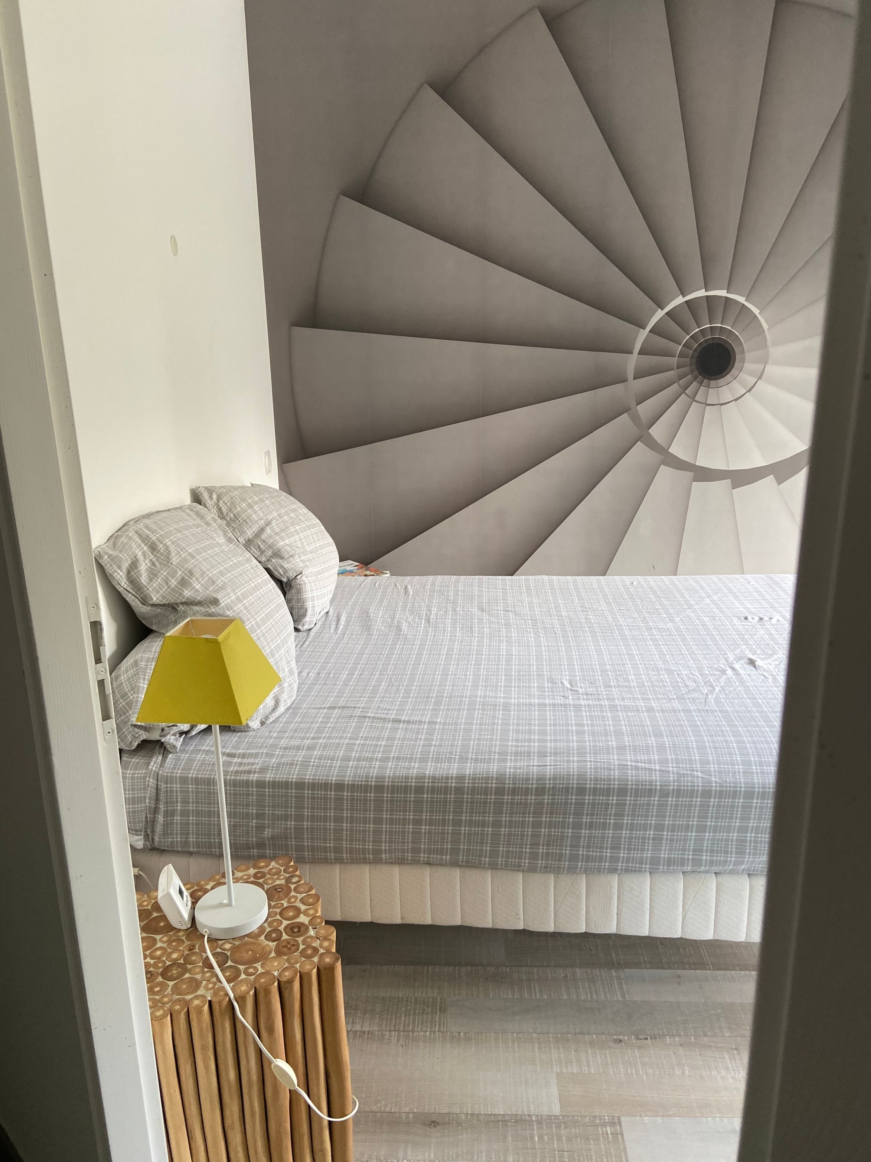 Une chambre avec un escalier en colimaçon, un lit avec une literie à carreaux, une lampe de sol jaune et une table de chevet avec une lampe.