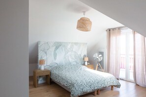 Une chambre avec un grand lit, une lampe fixée au mur, un ventilateur sur pied et une fenêtre avec des rideaux.