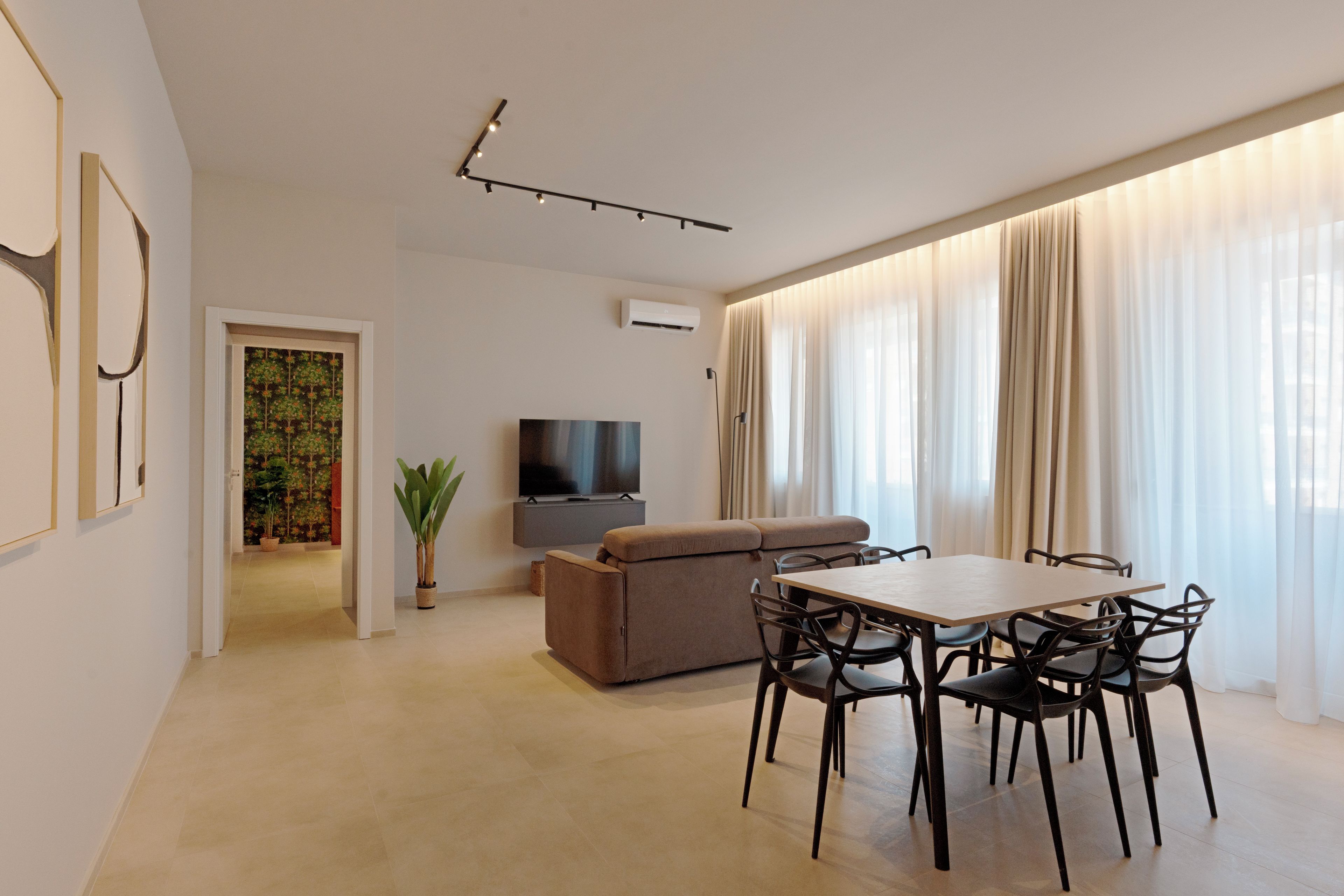 A modern living room with a flat-screen TV, a sofa, a dining table with chairs, and a large window with sheer curtains.