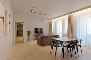 Una sala de estar moderna con un televisor de pantalla plana, un sofá, una mesa de comedor con sillas y un ventanal con cortinas translúcidas.