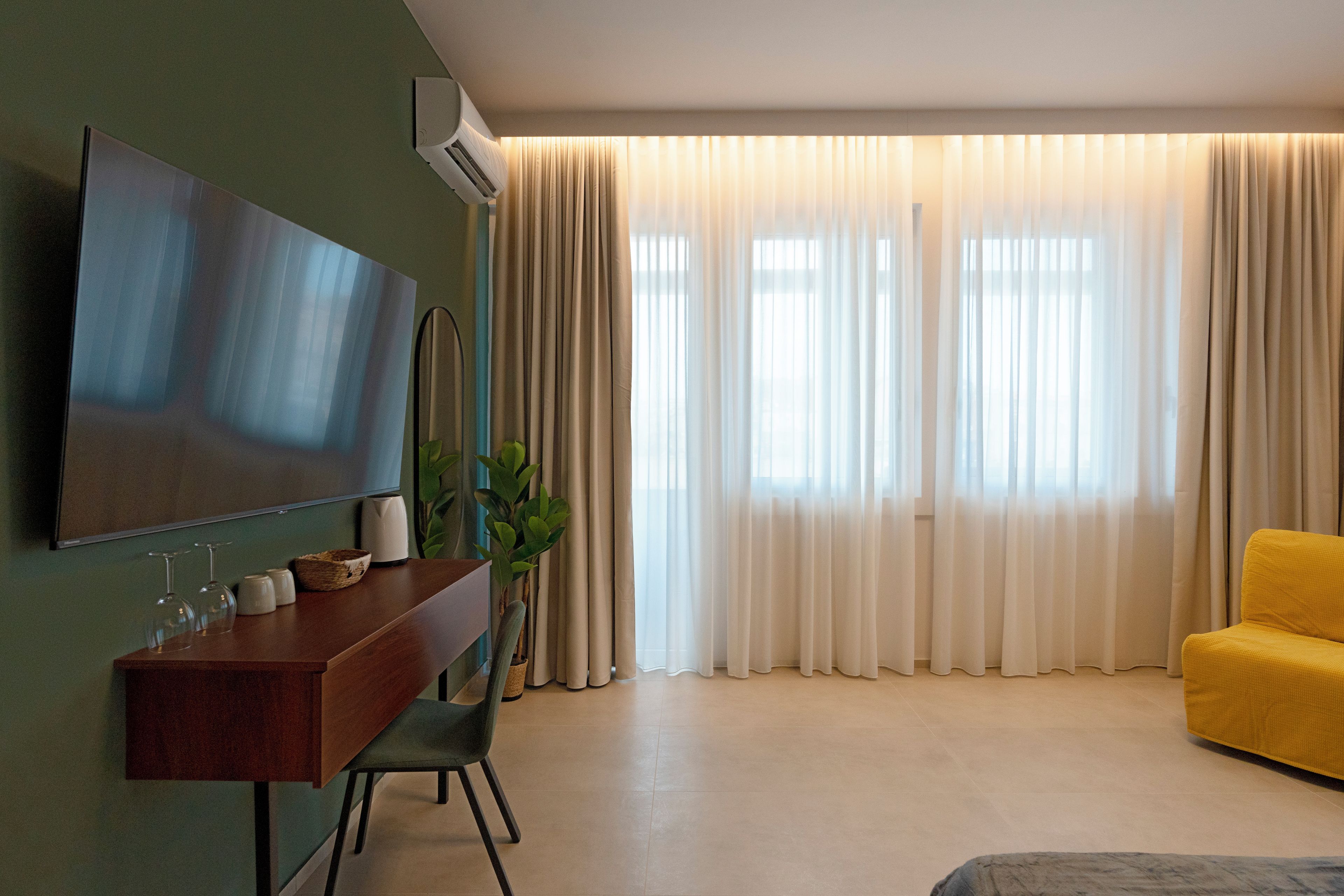 Una sala de estar moderna con un televisor de pantalla plana, un escritorio de madera, un sofá amarillo y un ventanal con cortinas translúcidas.