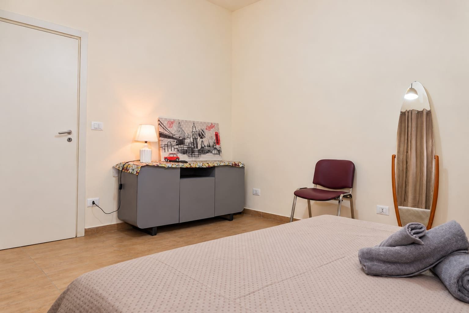 A bedroom with a bed, a desk with a lamp, a chair, a mirror, and a painting on the wall.