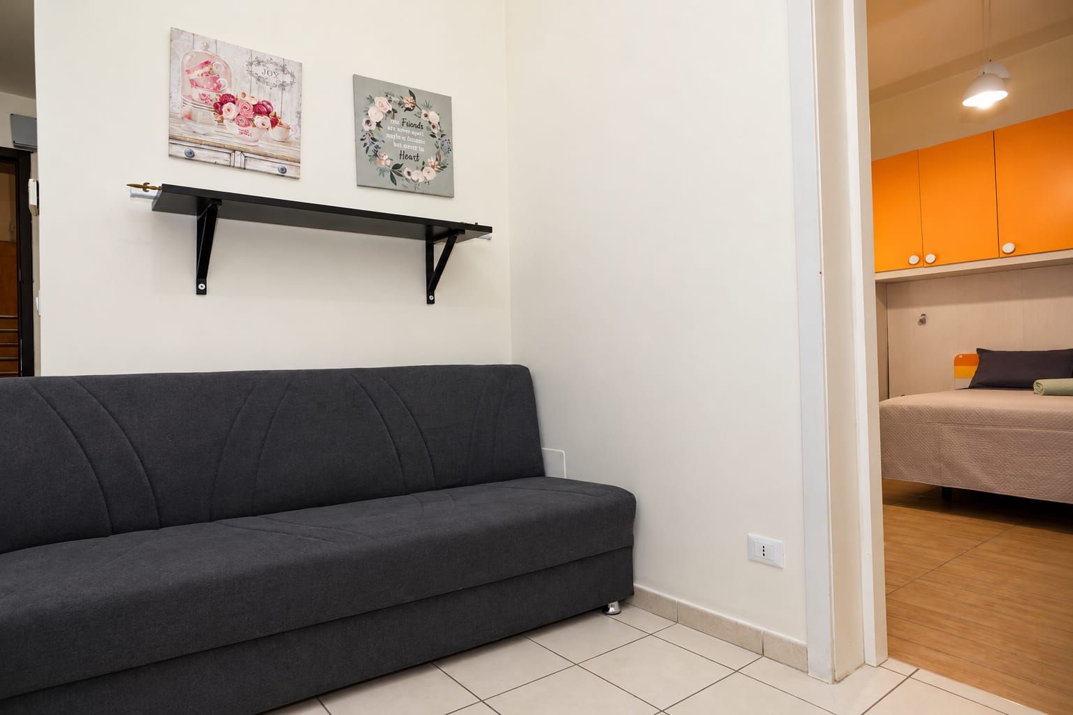 Una habitación con un sofá gris, una cama con colchón blanco y un estante de pared con dos cuadros enmarcados.
