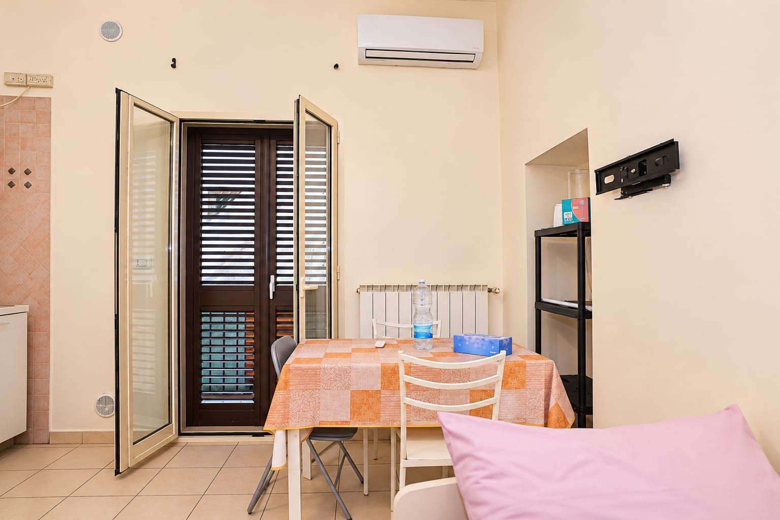 A compact room with a table, chairs, a water bottle, a box, a pink pillow, a radiator, an air conditioner, and a sliding door.