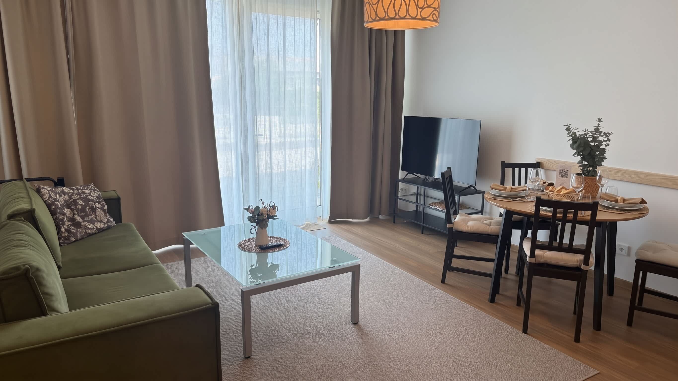 Ein Wohnzimmer mit einem grünen Sofa, einem Glastisch, einem Essbereich mit Tisch für zwei Personen, einem Fernseher und einer Pflanze.