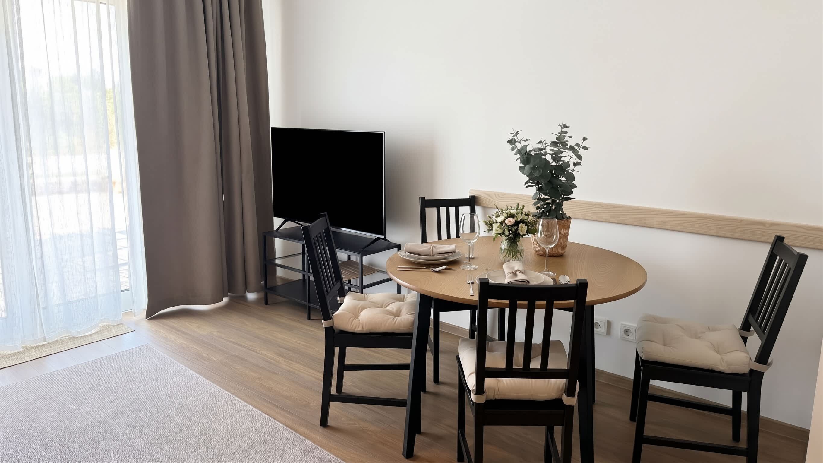 Ein Essbereich mit einem runden Tisch, schwarzen Stühlen mit Kissen, einer Zimmerpflanze und einem Fernseher auf einem Schrank.