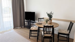 Ein Essbereich mit einem runden Tisch, schwarzen Stühlen mit Kissen, einer Zimmerpflanze und einem Fernseher auf einem Schrank.