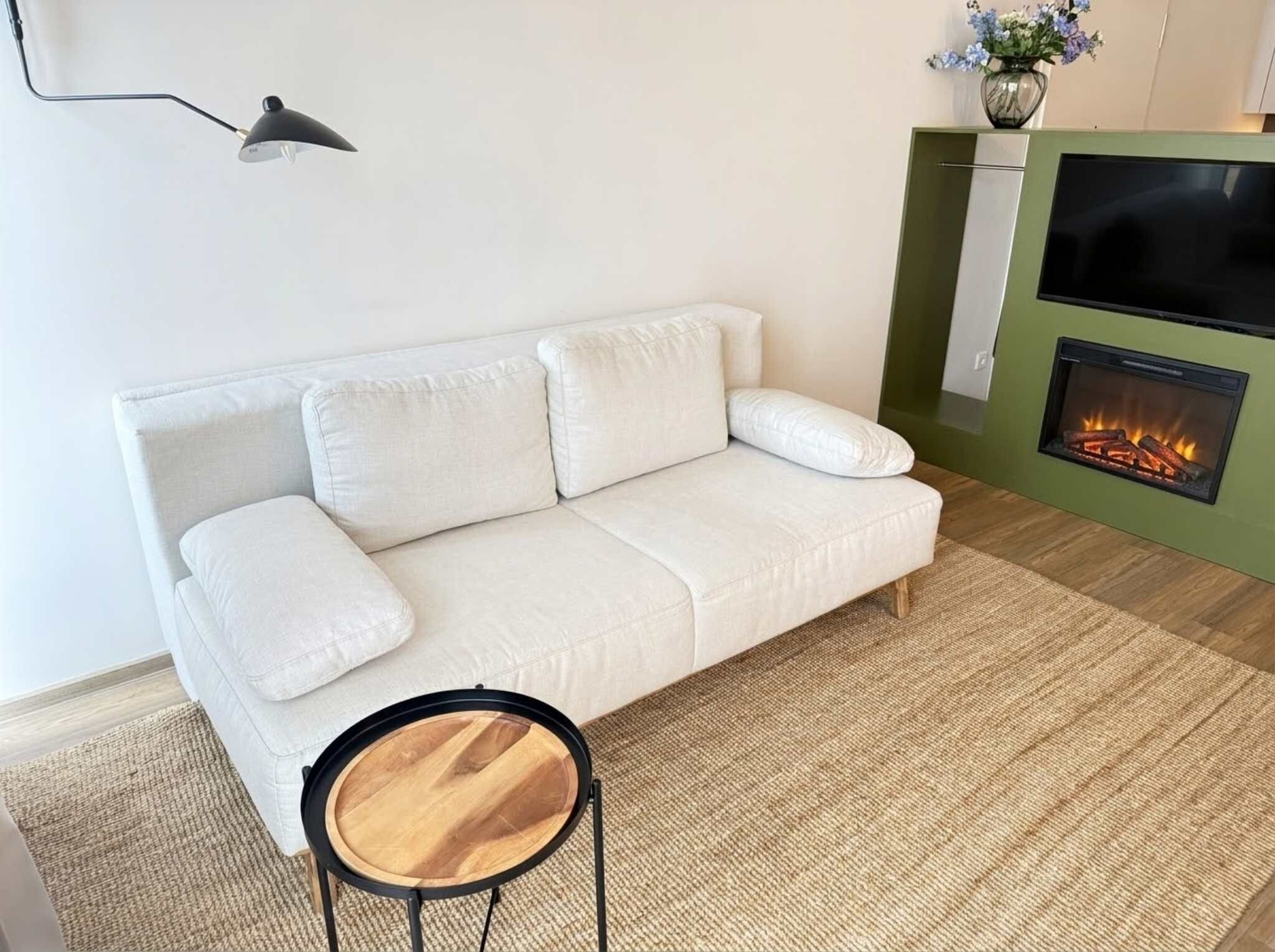 Ein weißes Sofa mit Kissen, ein kleiner runder Holztisch, eine schwarze Stehlampe und eine grüne Wand mit Kamin.