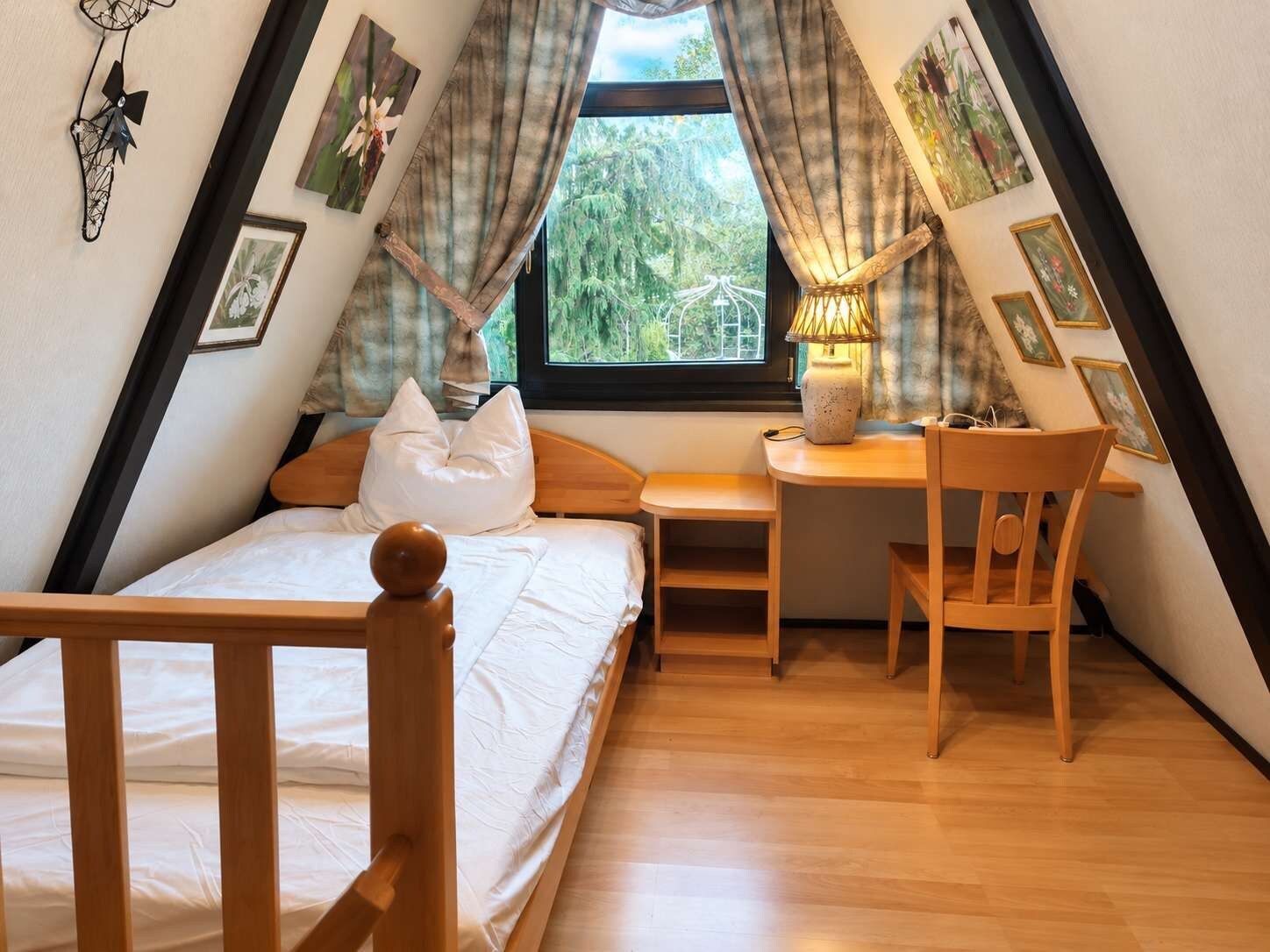 Ein Schlafzimmer mit Bett, Schreibtisch, Stuhl, einem Fenster mit Vorhängen und Blick ins Grüne.