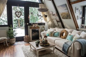 Ein gemütliches Wohnzimmer mit Kamin, einer Couch mit Kissen, einem Couchtisch und Blick ins Freie durch ein großes Fenster.