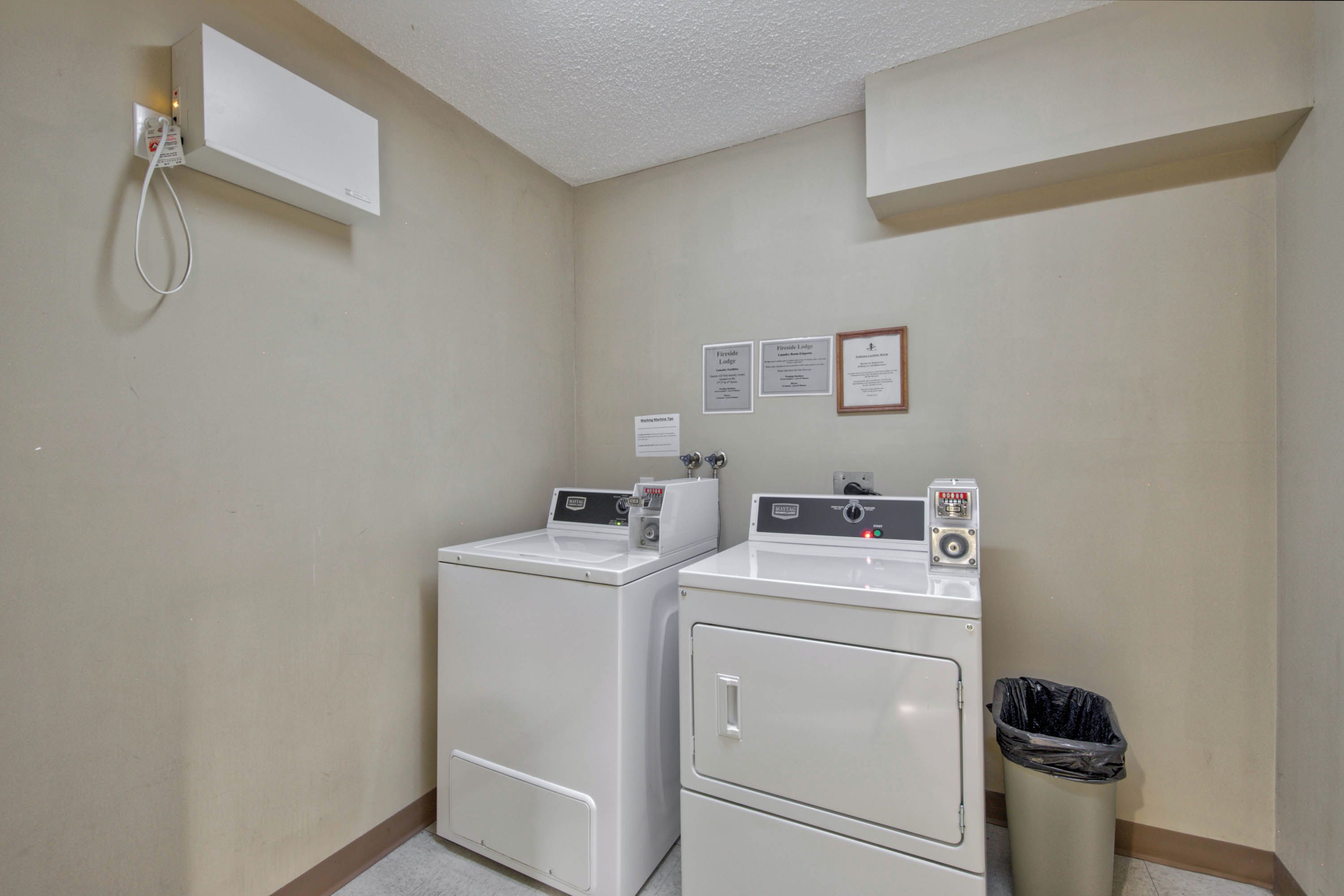 A laundry room with two stacked washers and dryers, a trash can, and a wall-mounted electrical outlet.