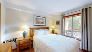 A hotel room with a large bed, two bedside tables with lamps, a painting on the wall, and a window with curtains. - Fireside #202 | 3 Peaks Rentals (Sun Peaks)