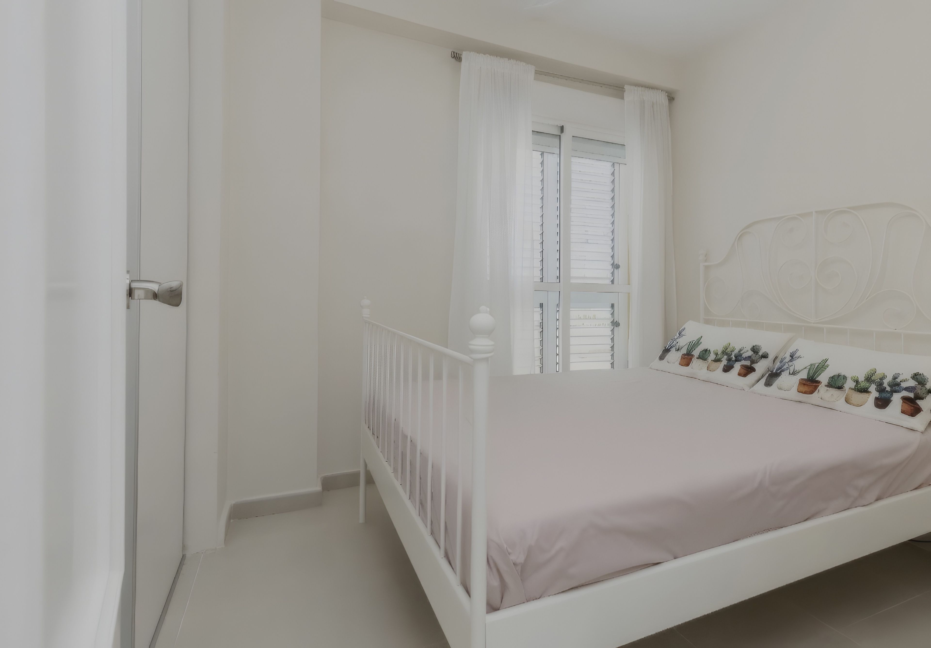 Een slaapkamer met een wit bed, een raam met jaloezieën en een deur.