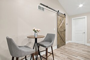 A room with a wooden floor, two grey chairs, a small round table with a vase of flowers, a sliding barn door, and a white door.