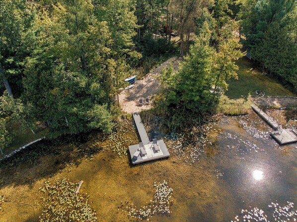An aerial view of a lakeside property with a dock, a small boat, and a picnic area surrounded by dense trees. - Lakefront Retreat | Sandy Beach, Private Dock, Kayaks, Fire Pit & Arcade Games (Gladwin)