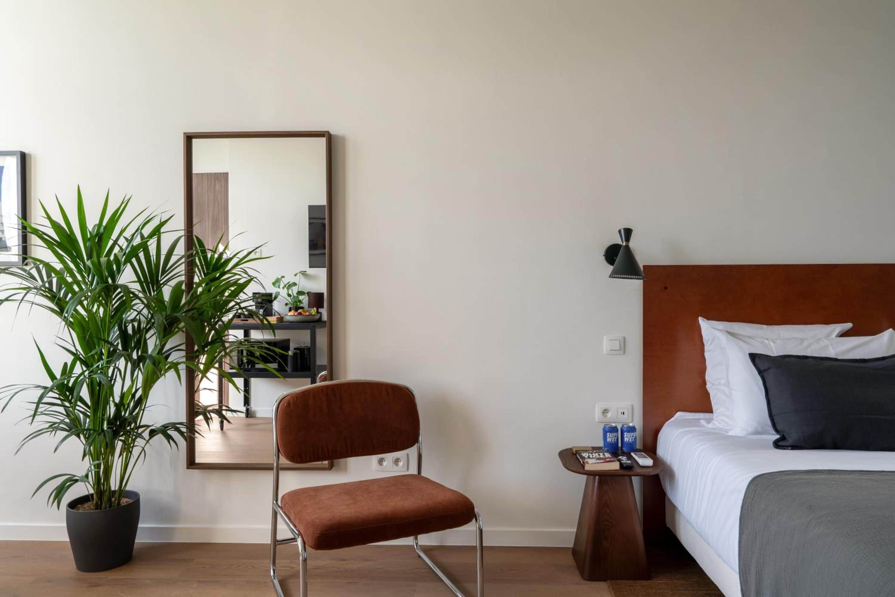 Quarto de hotel com uma cama, uma cadeira, uma mesa pequena, mesa de cabeceira, abajur e uma planta em vaso.