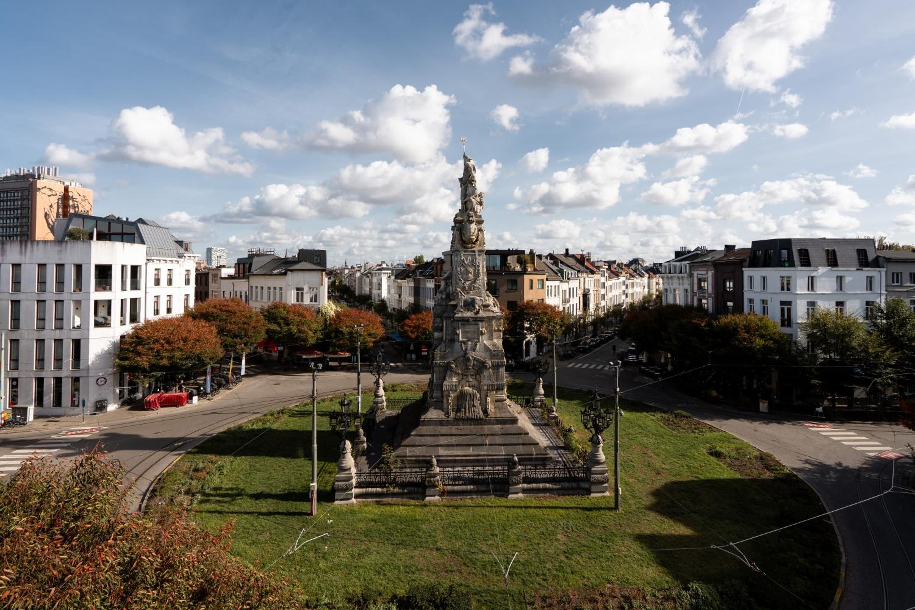 Eine Stadtansicht mit einem zentralen Denkmal, umgeben von einer Grünfläche, flankiert von Gebäuden.
