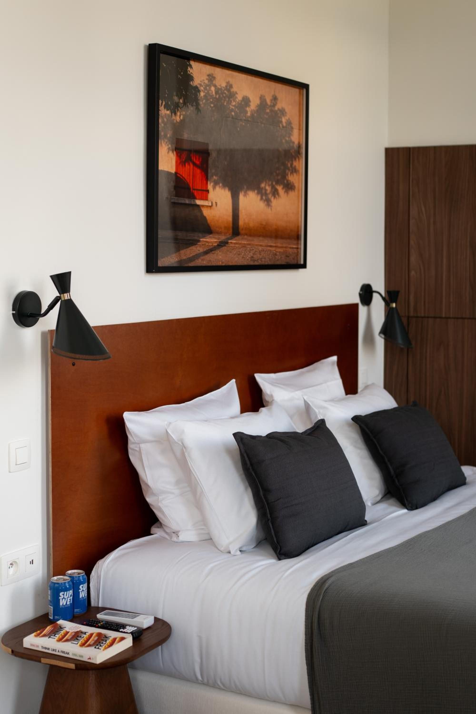 Ein ordentlich bezogenes Bett mit weißen und dunklen Kissen, ein gerahmtes Bild darüber und je eine Wandlampe an jeder Seite.