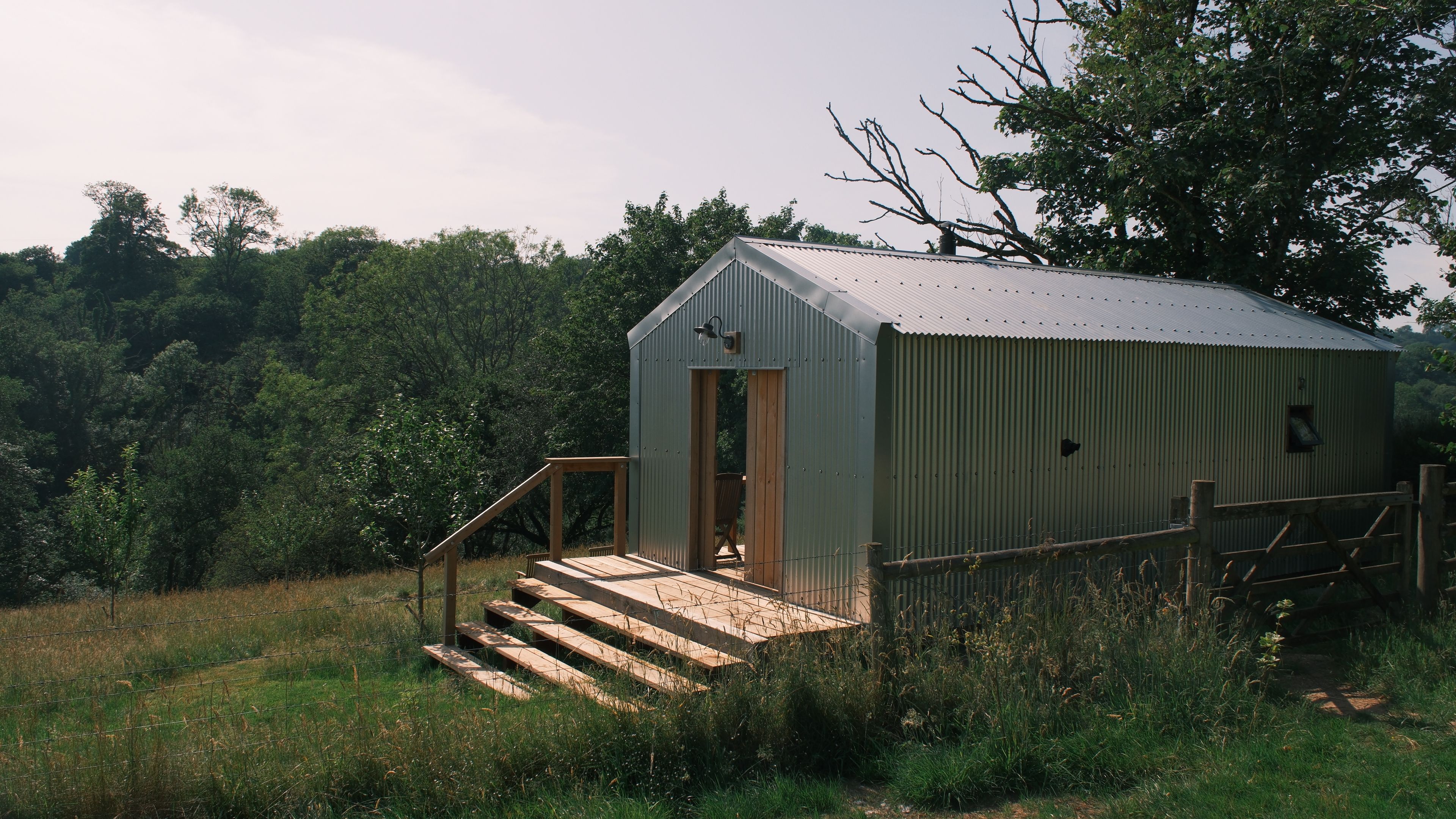 A metal shed with a wooden door and steps leading up to it, surrounded by grass and trees.