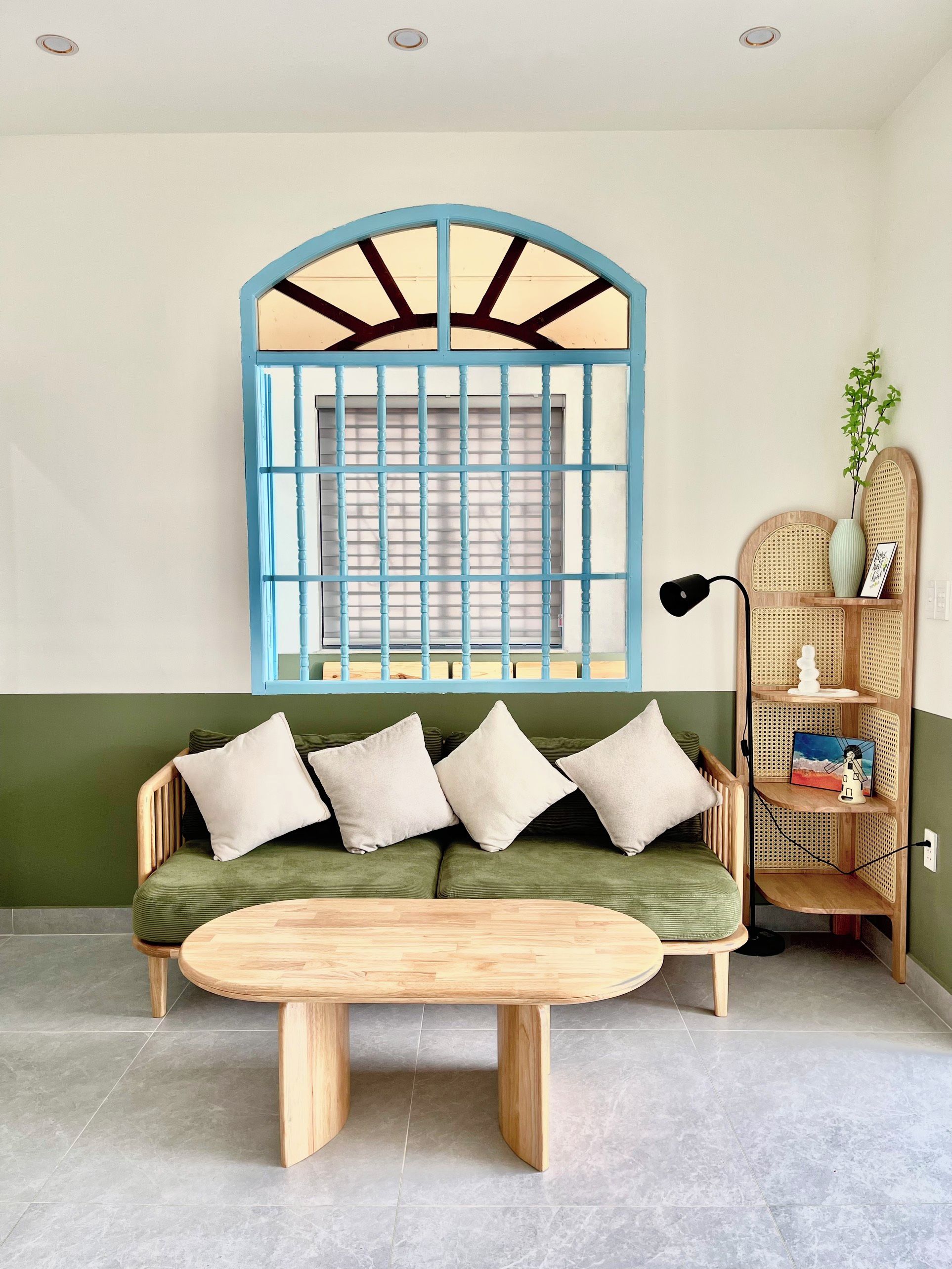 Una habitación con un sofá verde, una mesa de madera, un ventanal con marco azul y un estante de mimbre con adornos.