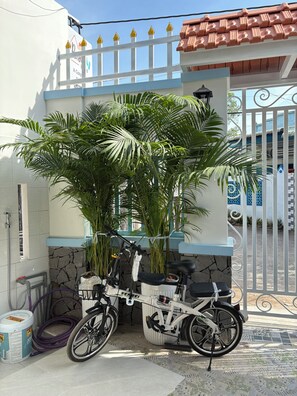 Uma bicicleta branca estacionada em frente a uma casa com portão azul e branco, rodeada por vegetação.