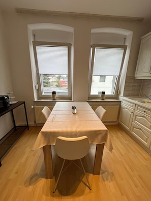 Et køkken med spisebord, to vinduer med rullegardiner, en radiator og en mikrobølgeovn på en hylde.