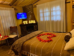 Una camera d'albergo con camino in pietra, un letto decorato con una ghirlanda a forma di cuore, una televisione e una poltrona.