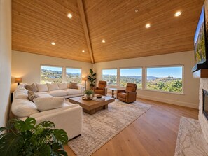 A spacious living room with a high ceiling, large windows offering a view of the countryside, a fireplace, and a comfortable seating area. - Ocean-View Villa Jacuzzi Near Laguna Seca (Monterey)