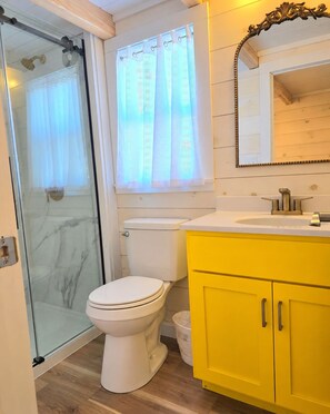 A bathroom with a glass shower enclosure, a toilet, a sink with a mirror, and yellow cabinets.