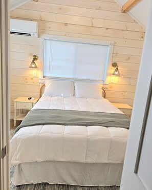 A bedroom with a bed, two bedside tables, a window with blinds, and wall-mounted lamps.