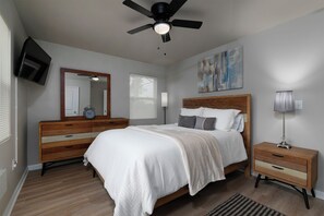 Quarto com cama, mesa de cabeceira, cômoda, ventilador de teto, televisão e janela com persianas.