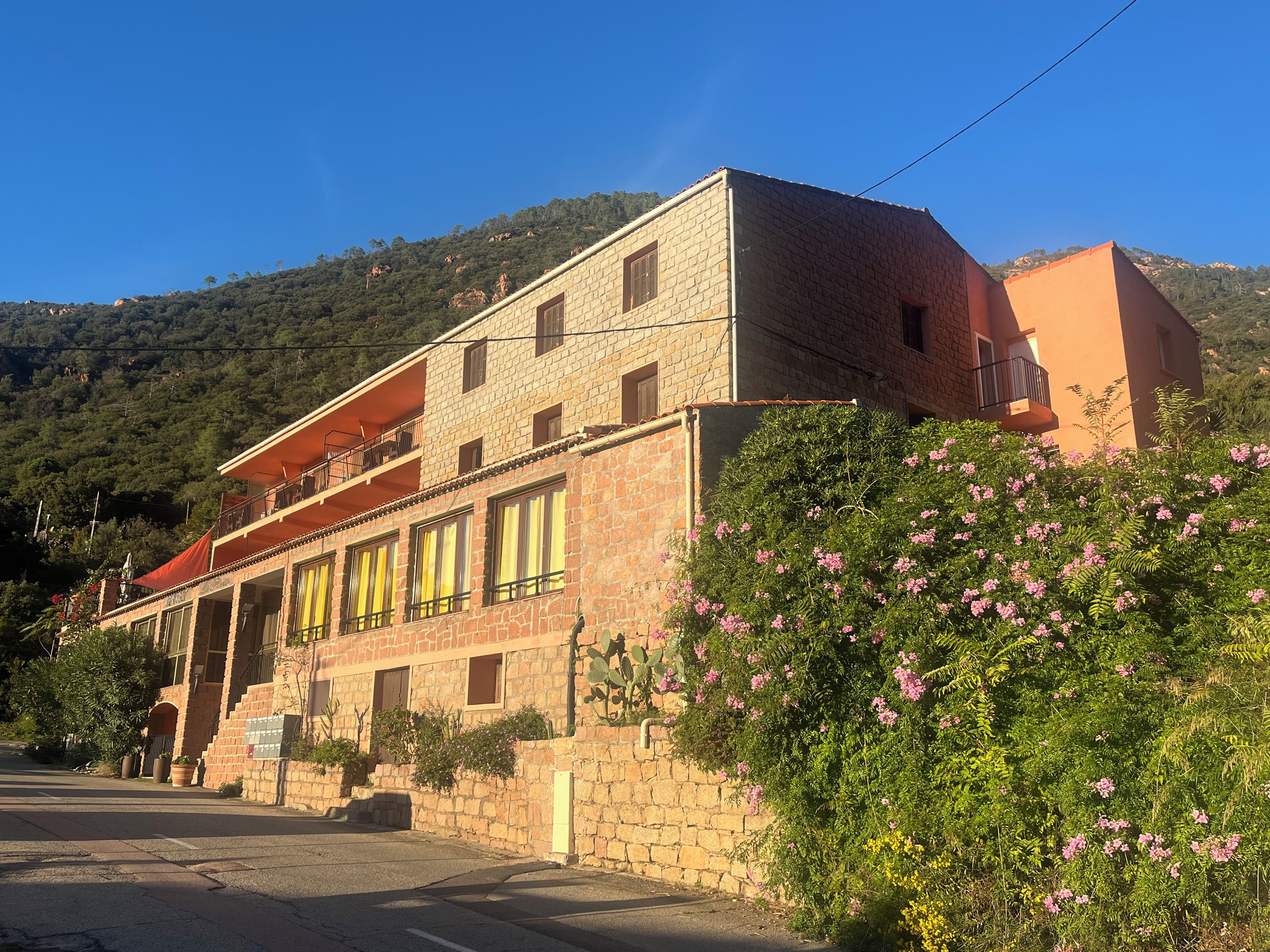 A two-story stone building with a balcony, surrounded by greenery and hills.