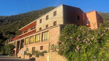 A two-story stone building with a balcony, surrounded by greenery and hills.