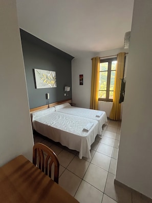 A hotel room with two beds, a dining table, a chair, a window with curtains, and a wall-mounted light. - New Listing (Ota)