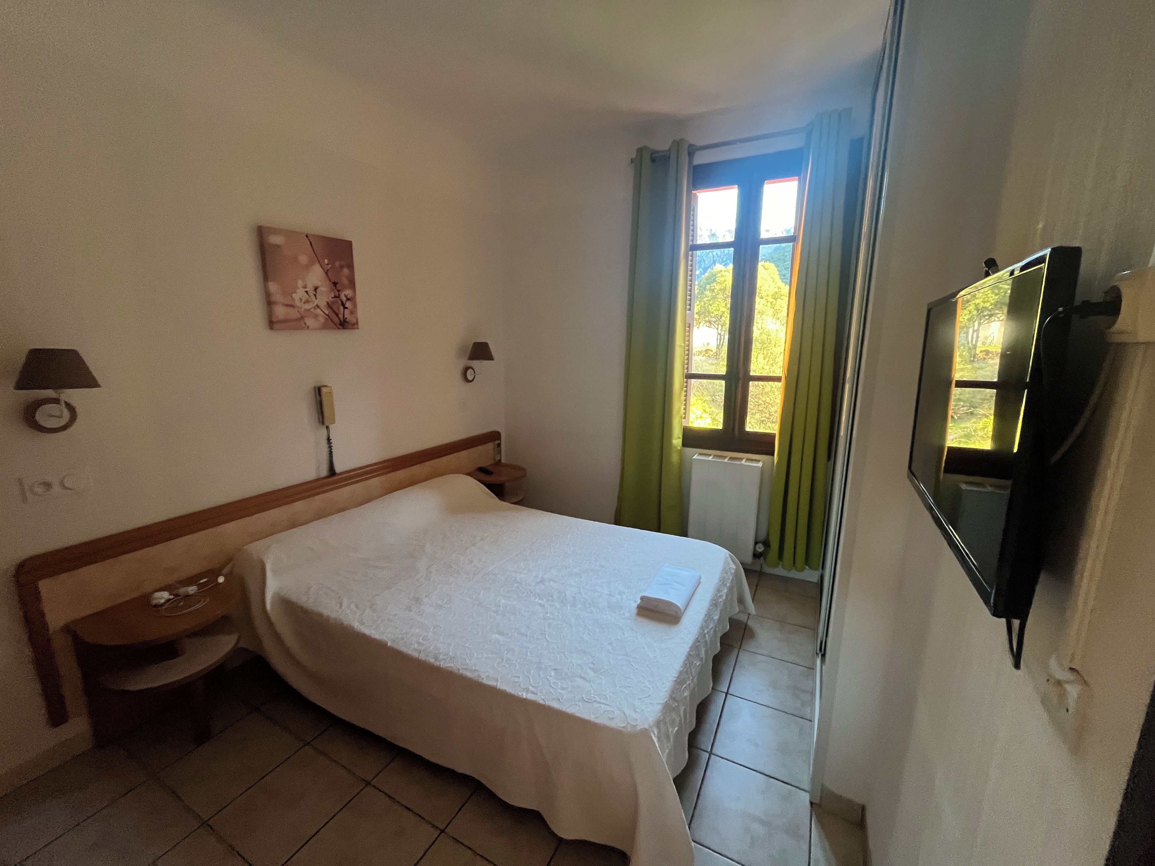 Una habitación de hotel con cama, mesita de noche, lámpara, reloj de pared y televisor.