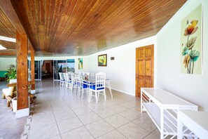 Un comedor amplio con sillas blancas y una mesa larga, vigas de madera en el techo y una puerta de madera.