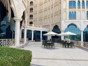Un espace extérieur aménagé en terrasse, avec des tables et des chaises, un bar, et un bâtiment doté de fenêtres en arc de cercle et de balcons.