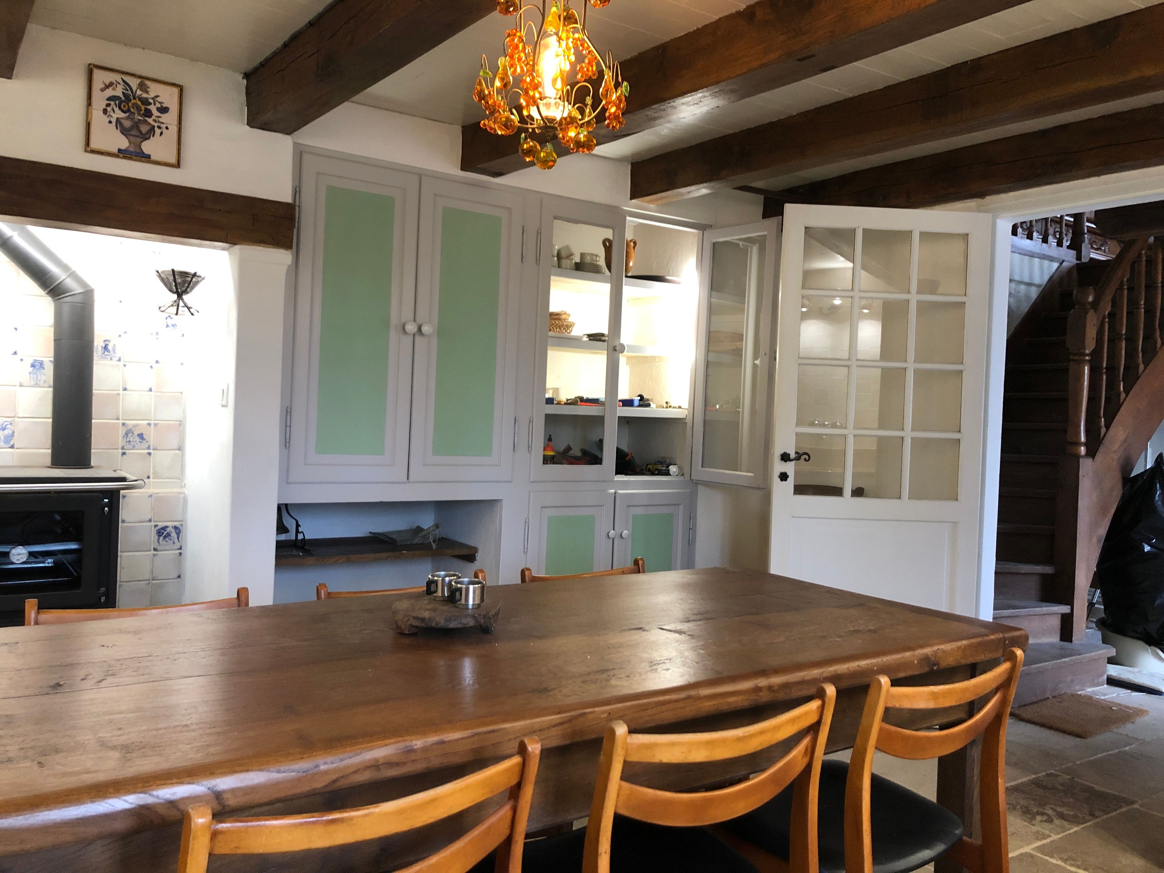 Uma área de refeições com mesa e cadeiras de madeira, uma cozinha com armários brancos e uma escada que leva a outro andar.